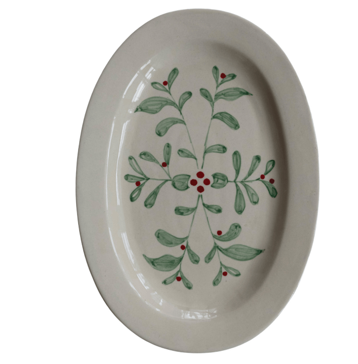 An oval ceramic serving platter with a glossy cream glaze, decorated with a hand-painted green mistletoe and red berries motif in the center.