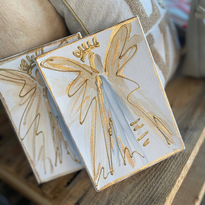 Two square white canvas art prints featuring gold and light blue angel designs, displayed on a rustic wooden shelf next to neutral-toned cushions.