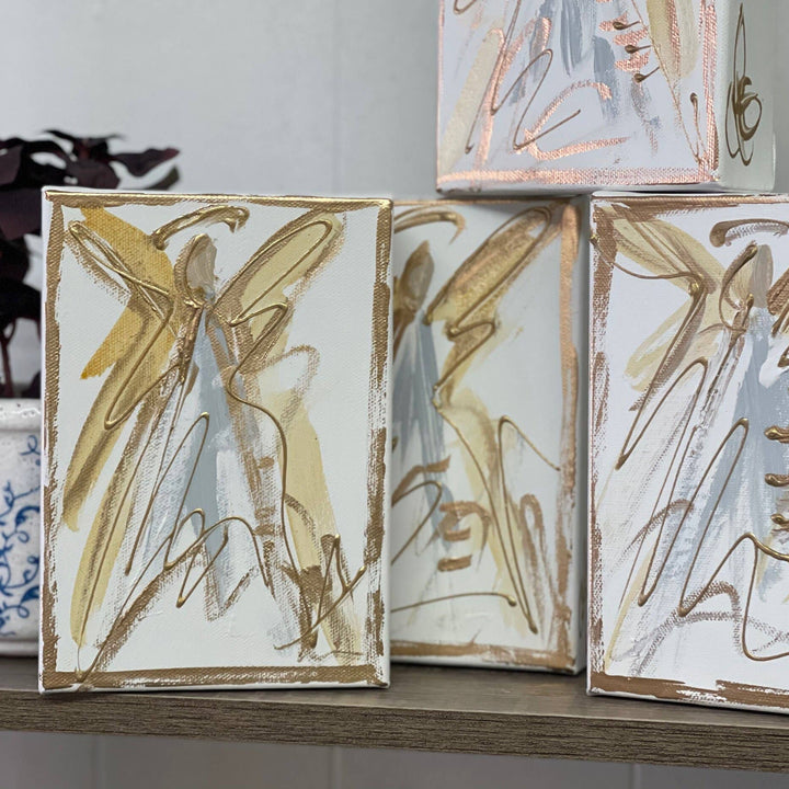 Three small abstract art canvases with gold and gray brushstrokes on a white background, displayed on a wooden shelf next to a potted plant.