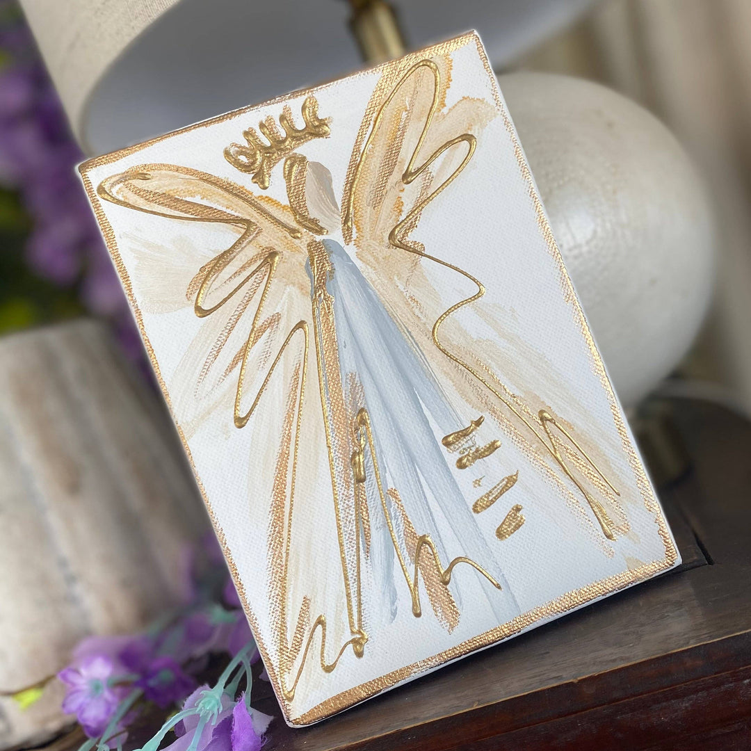 A square decorative wall art piece featuring a stylized angel figure in gold and white tones, displayed leaning on a dark wooden table next to a lamp.