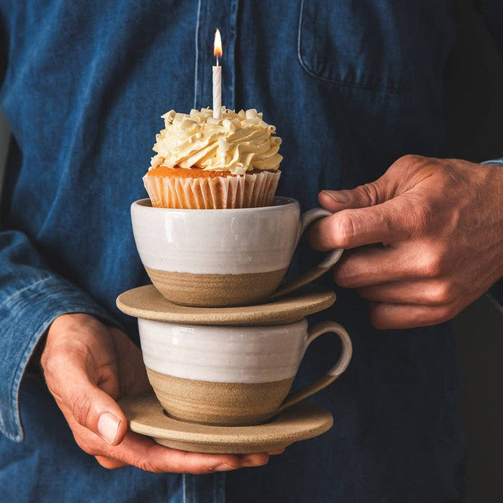 stack of two Pantry Mugs with saucer and a cupcake with a candle in the top one and being held by someone wearing a blue denim shirt