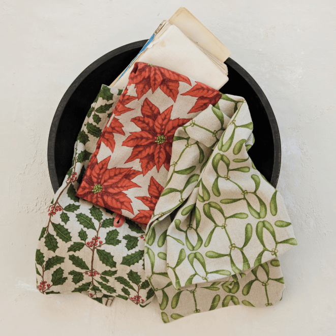 Three folded patterned fabric napkins or cloths in a black bowl on a light textured background. One cloth has red poinsettias on cream, one has green mistletoe on cream, and one has green holly leaves with small red berries on cream.