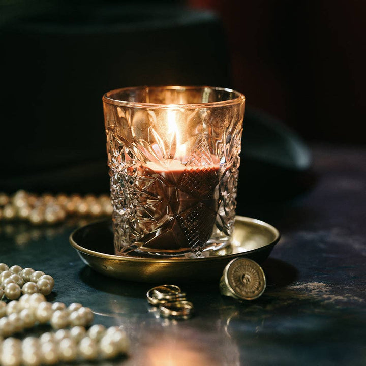 Candle in a textured glass holder on a dark surface with pearls and coins.