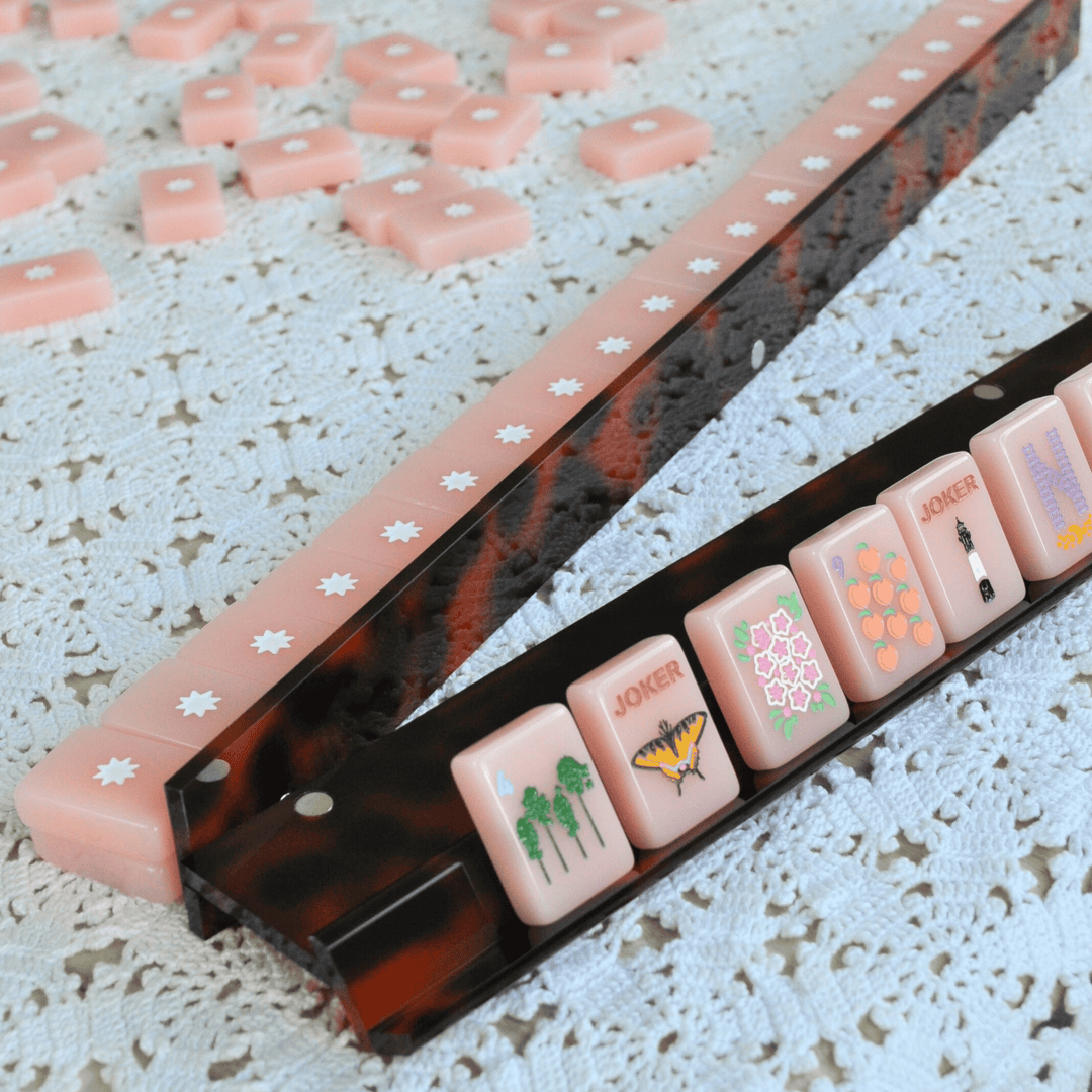 Tortoise shell mahjong rack and pusher with pink mahjong tiles on a white tablecloth.