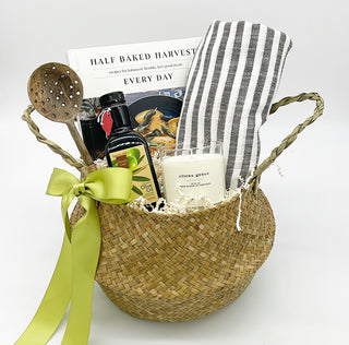 Gift for the gourmet includes Half Baked Harvest book, wooden olive spoon, olive oil and citrus scented candle in a rattan belly basket tied with satin or grosgrain ribbon