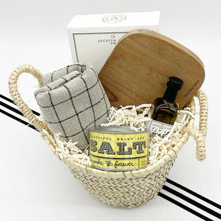 Petite Kitchen Housewarming Basket is filled with traditional housewarming gifts pulled together in a reusable rattan basket. Items include artisanal crackers, olive oil, flavored salt, kitchen towel, cutting board. 